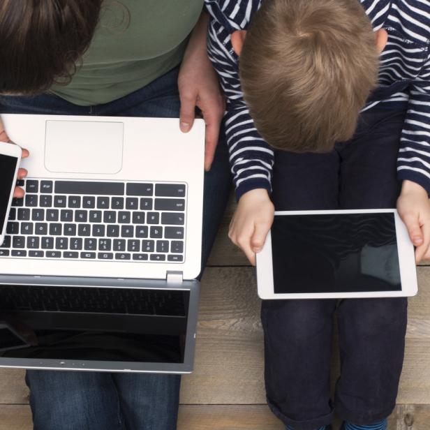 Eine Frau mit Laptop und Handy sitzt neben einem Kind mit Tablet auf einem Holzboden.
