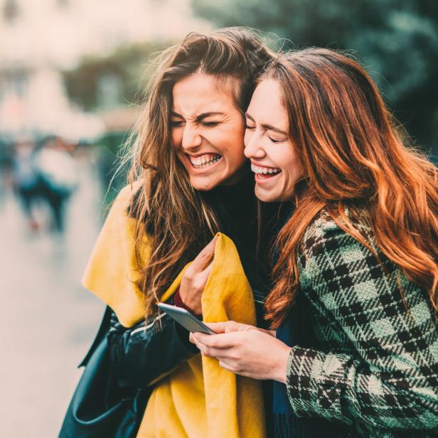 Zwei junge Frauen umarmen sich und lachen, während sie auf ein Smartphone schauen.