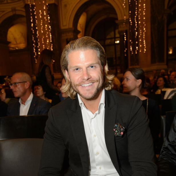 Ein lächelnder Paul Janke mit blonden Haaren sitzt im Anzug in einem Saal mit Publikum.