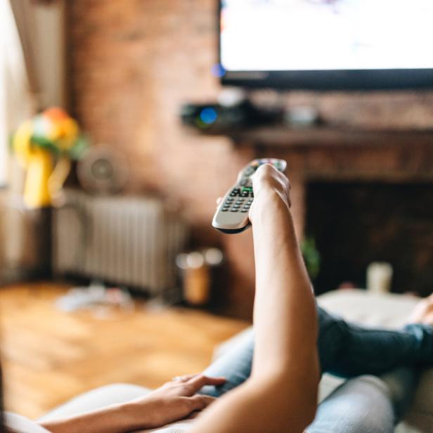 Eine Person liegt auf einem Sofa und zappt mit einer Fernbedienung durch das Fernsehprogramm.