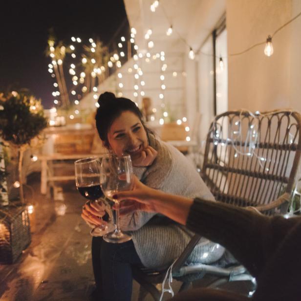 Eine Frau prostet mit einem Glas Rotwein auf einer mit Lichterketten dekorierten Terrasse zu.