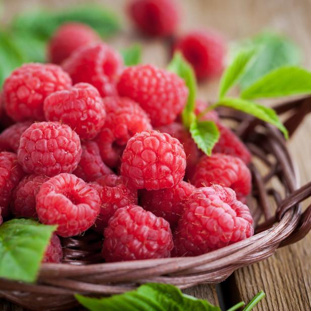 Ein Korb voller frischer Himbeeren mit grünen Blättern auf einem Holztisch.