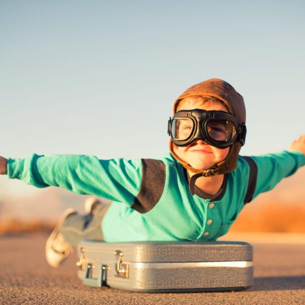 Ein Junge mit Fliegerbrille und -mütze liegt auf einem Koffer und tut so, als würde er fliegen.