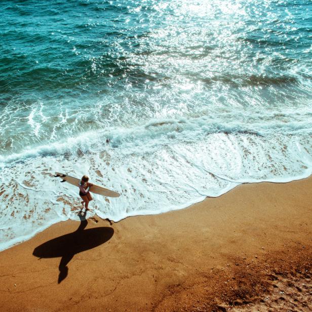 Eine Frau mit Surfbrett steht am Strand im türkisfarbenen Wasser.