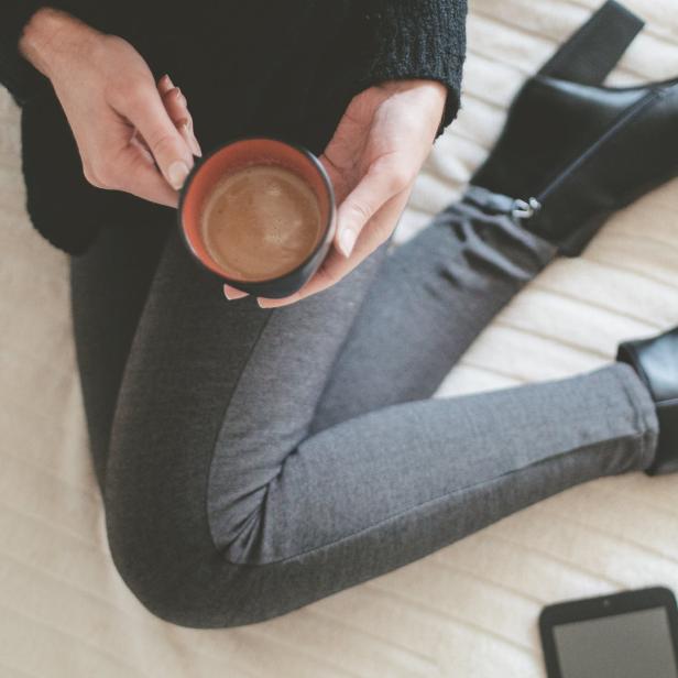 Eine Frau sitzt auf einem Bett, hält eine Tasse Kaffee und trägt schwarze Stiefel.