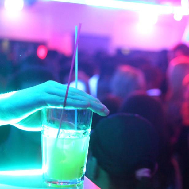 Ein Trinkglas mit einer Hand darauf, im Hintergrund eine Szene aus einem Tanzklub