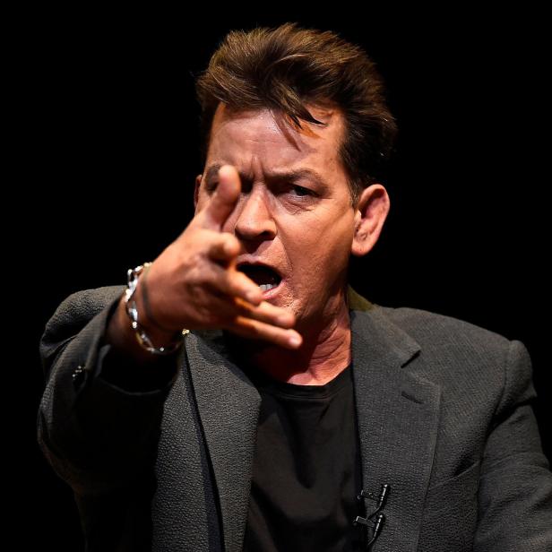 Charlie Sheen gestikuliert mit erhobener Hand vor einem schwarzen Hintergrund.