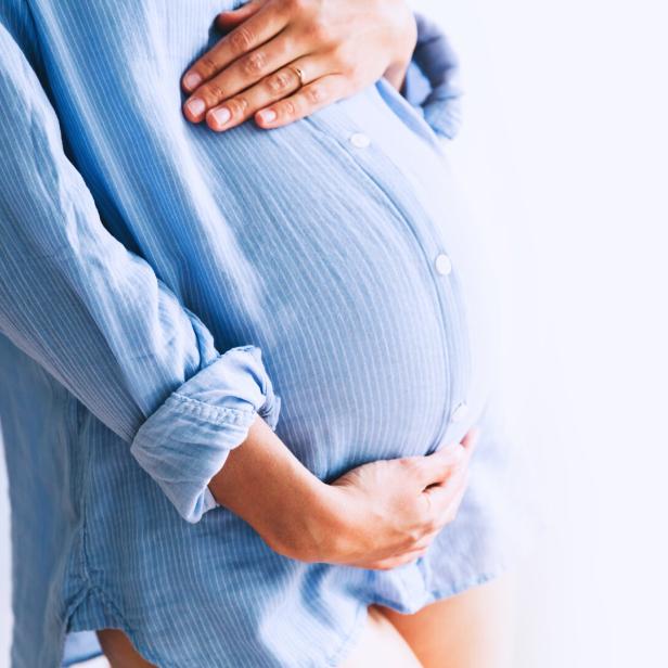 Eine schwangere Frau in einem hellblauen Hemd hält ihren Bauch.