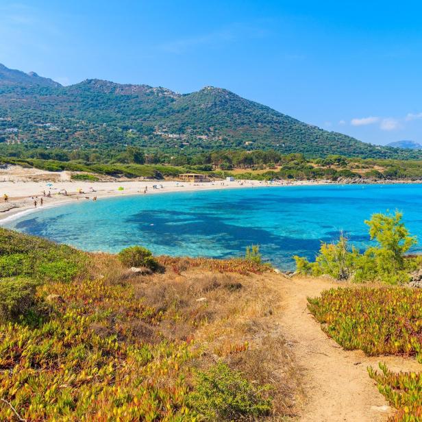 Ein Sandweg führt zu einem Strand mit türkisfarbenem Wasser und Bergen im Hintergrund.