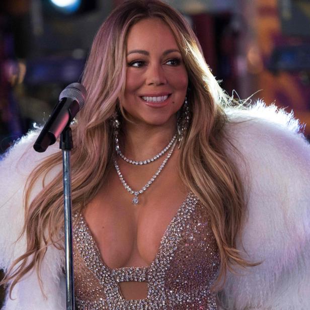 Mariah Carey steht lächelnd mit Glitzerkleid und weißem Fellmantel vor einem Mikrofonständer.