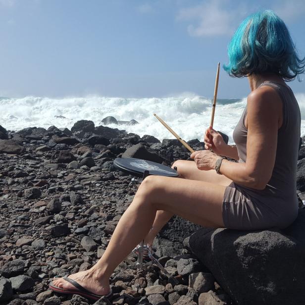 Eine Frau mit blau gefärbten Haaren sitzt am Strand und spielt Schlagzeug.