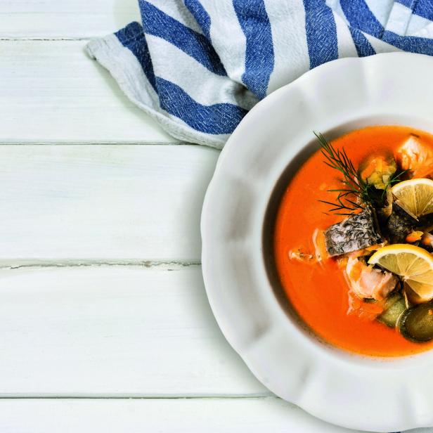 Eine Schüssel Fischsuppe mit Zitrone und Dill auf einem Tisch mit blau-weiß gestreiftem Tuch.