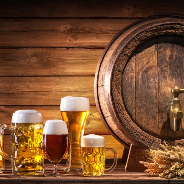 Verschiedene Biersorten stehen vor einem Holzfass mit Hopfen und Weizenähren.