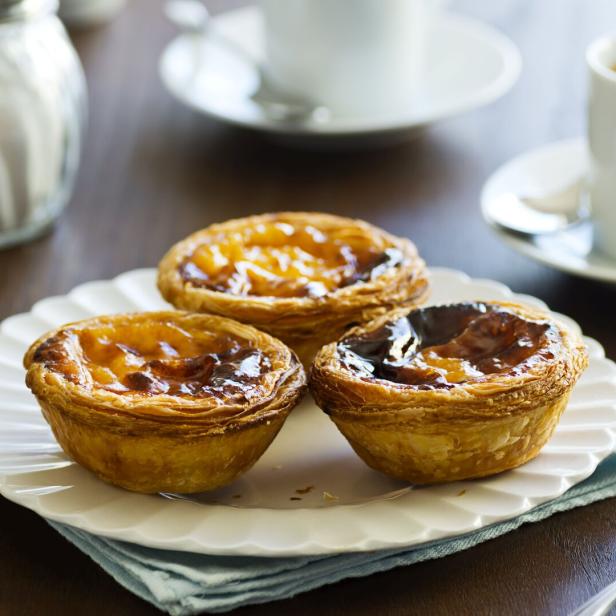 Drei Pastéis de Nata auf einem Teller mit Tassen im Hintergrund.