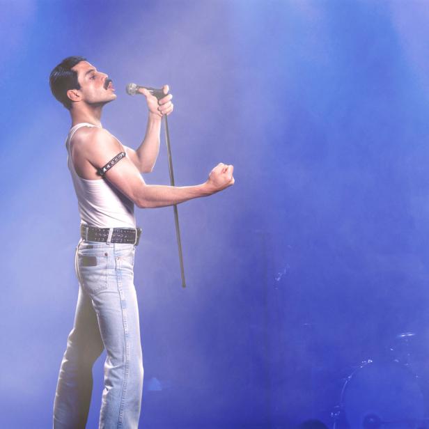 Ein Mann im Stil von Freddie Mercury singt mit einem Mikrofon vor blauem Hintergrund.