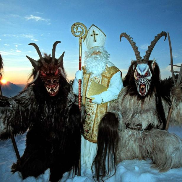 Ein Nikolaus steht mit zwei Krampus-Gestalten in einer verschneiten Landschaft.