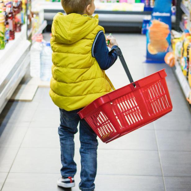 Ein Junge mit gelber Weste schiebt einen roten Einkaufswagen in einem Supermarkt.