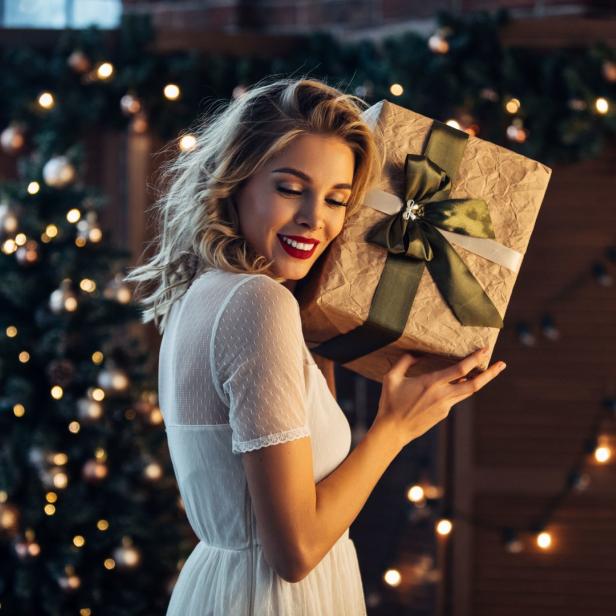Eine Frau hält freudig ein Geschenk vor einem festlich geschmückten Weihnachtsbaum.