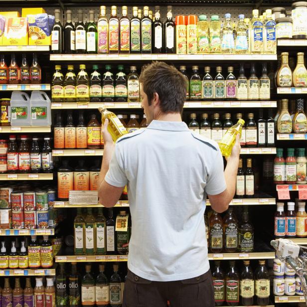 Ein Mann steht vor einem Regal im Supermarkt und vergleicht zwei Flaschen Öl.