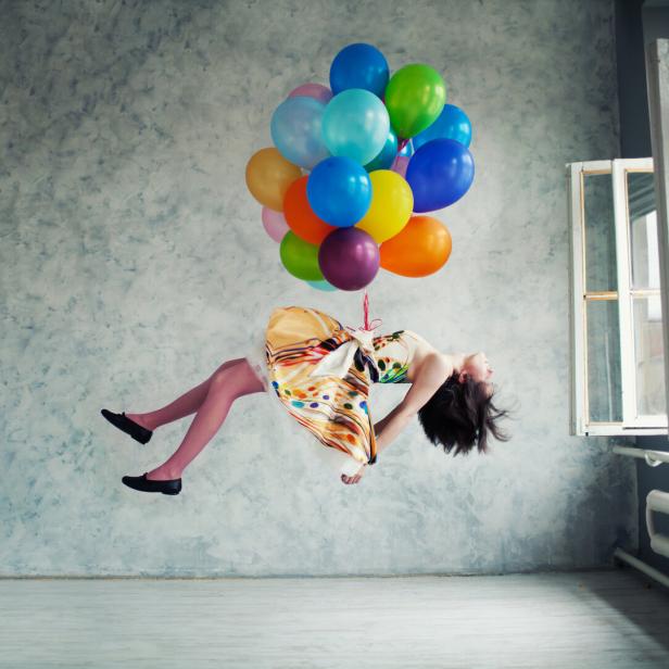 Eine Frau schwebt, an bunten Ballons befestigt, in einem Raum.
