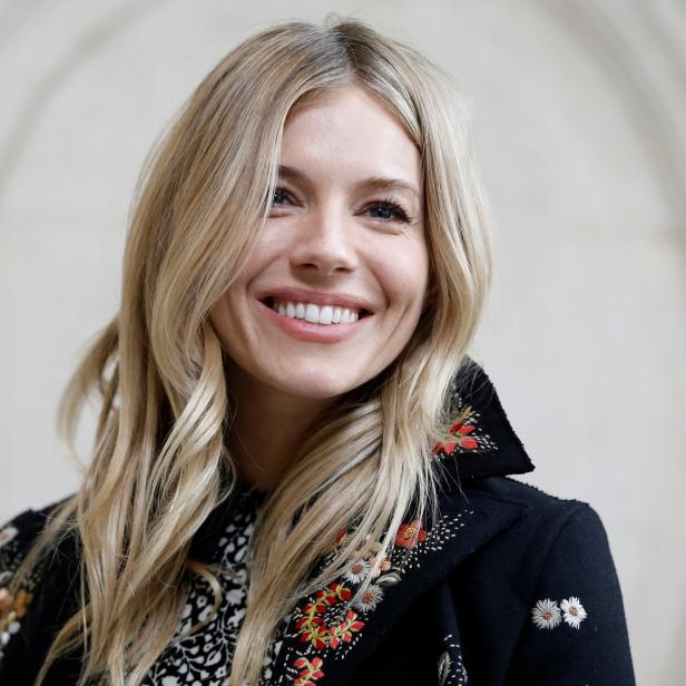 Sienna Miller lächelt in einem bestickten schwarzen Mantel.