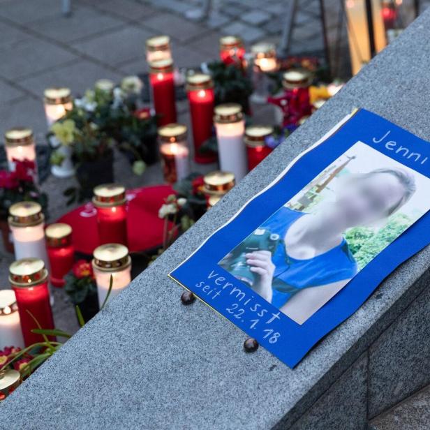 Ein Suchplakat für die vermisste Jenni liegt auf einer Steinmauer, umgeben von zahlreichen brennenden Grablichtern und Blumen.