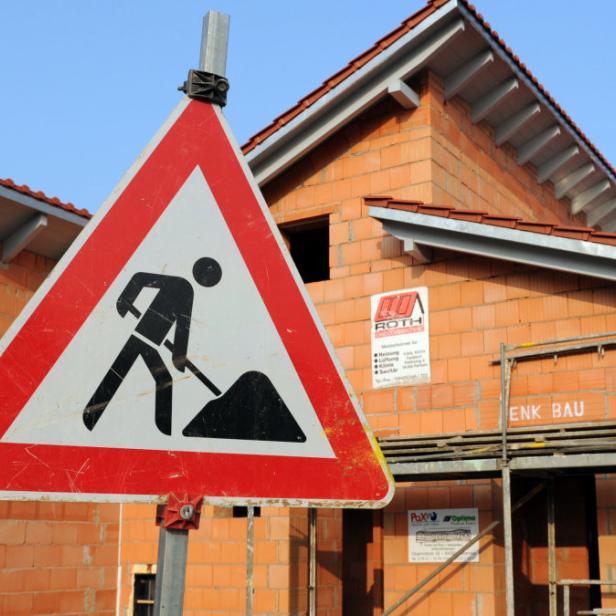 Ein Baustellenschild steht vor einem im Rohbau befindlichen Haus mit Gerüst und roten Dachziegeln.