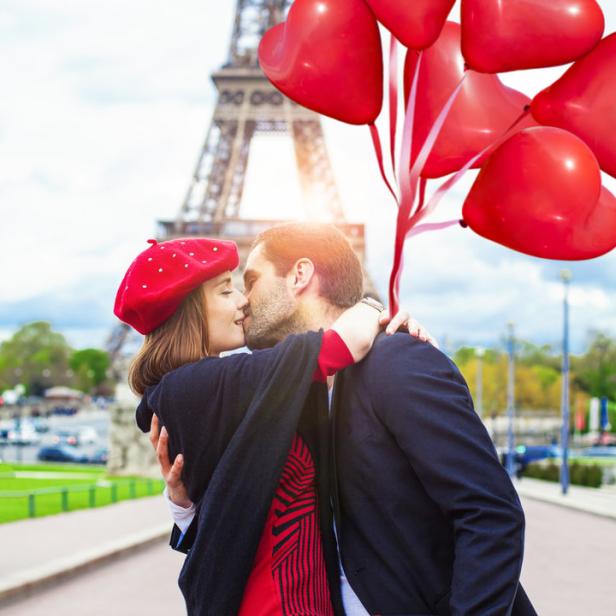 Ein Paar küsst sich vor dem Eiffelturm, während der Mann rote Herzluftballons hält.