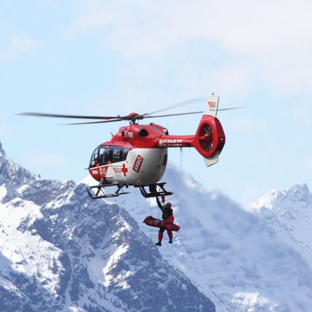 Ein Rettungshubschrauber fliegt vor verschneiten Bergen, während eine Person an einem Seil transportiert wird.