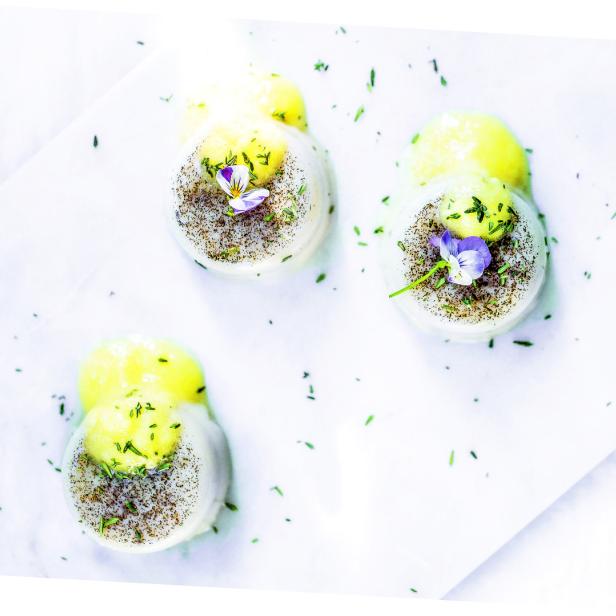 Drei Panna Cotta-Desserts mit gelbem Topping und essbaren Blüten auf einem weißen Tuch.