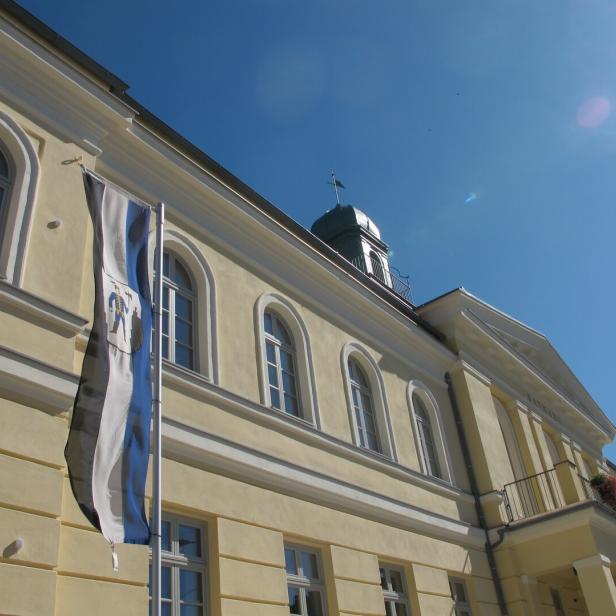 Helles Gebäude mit Rundbogenfenstern, Fahne mit Wappen und Balkon mit Blumenkästen unter blauem Himmel.