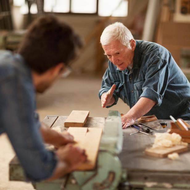 Ein älterer Mann erklärt einem jüngeren Mann in einer Werkstatt die Holzbearbeitung an einer Werkbank.