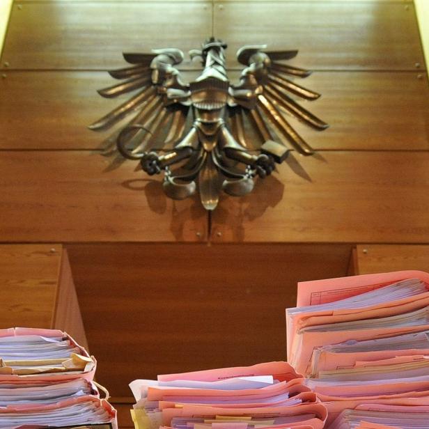 Eine Aufnahme aus einem Gerichtssaal, der den österreichischen Staatsadler auf einer Holzwand zeigt, darunter sind Zettel gestapelt.