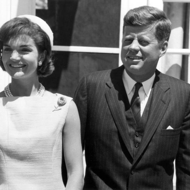 John F. Kennedy und Jacqueline Kennedy lächeln nebeneinander stehend vor einem hellen Gebäude.
