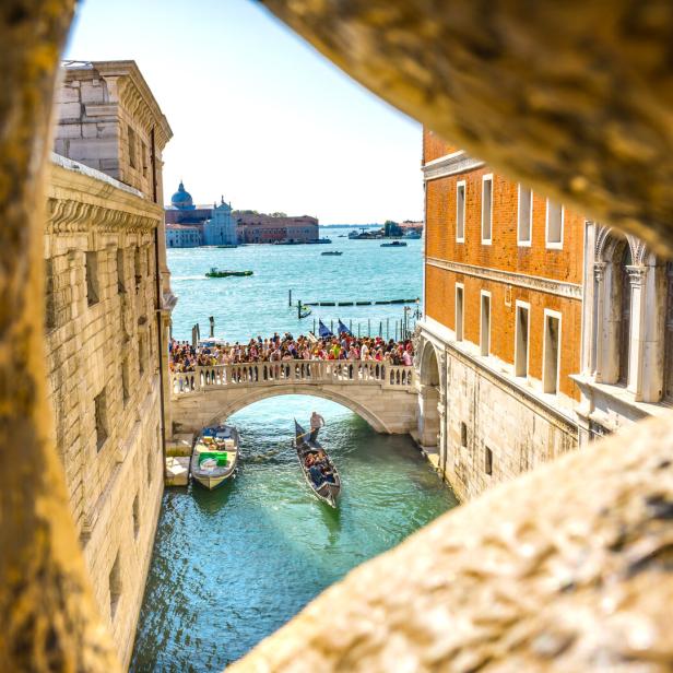 Blick durch einen steinernen Rahmen auf eine belebte Brücke und eine Gondel in Venedig.