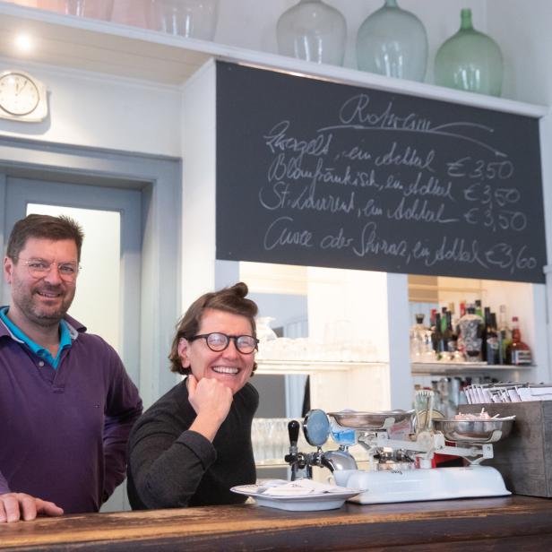 Zwei Personen stehen hinter einer Bar mit einer Tafel, die Weinsorten auflistet.