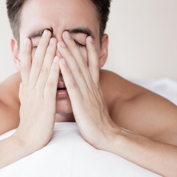 Ein Mann liegt im Bett und hält sich die Hände vors Gesicht.
