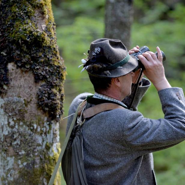 Ein Mann mit Trachtenhut beobachtet mit einem Fernglas die Umgebung im Wald.