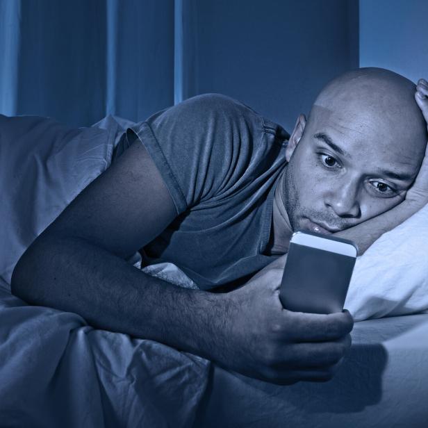 Ein Mann liegt nachts wach im Bett und schaut auf sein Handy.