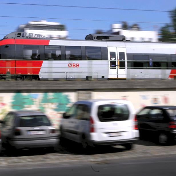 Ein roter und silberner ÖBB-Zug fährt an parkenden Autos vorbei.