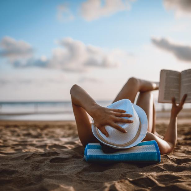 Eine Frau liest am Strand ein Buch, den Hut ins Gesicht gezogen.