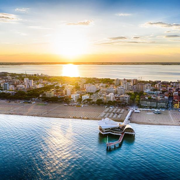 Luftaufnahme von einem Küstenort mit Strand, Meer und einem Restaurant auf einem Pier bei Sonnenuntergang.
