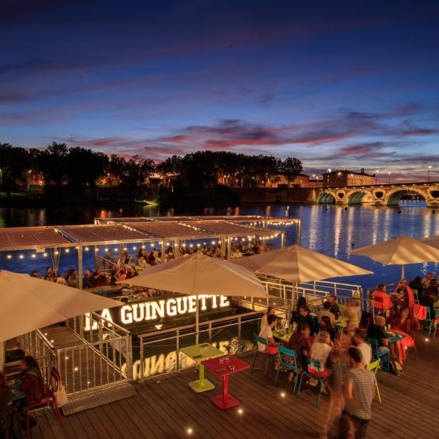 Das Restaurant „La Guinguette“ liegt auf dem Wasser, im Hintergrund eine Brücke bei Dämmerung.