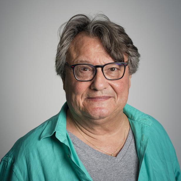 Ein Porträt eines Mannes mit Brille und einem grünen Hemd vor einem grauen Hintergrund.