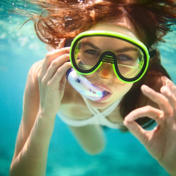 Eine Frau taucht mit Schnorchel und Taucherbrille im türkisfarbenen Wasser und zeigt ein OK-Zeichen.