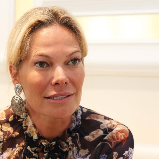 Eine blonde Frau mit Ohrringen und einer geblümten Bluse blickt zur Seite.