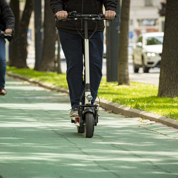 Eine Person fährt mit einem E-Scooter und eine weitere mit dem Fahrrad auf einem grünen Radweg entlang einer Baumallee.