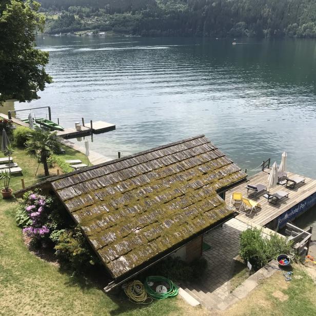 Blick auf einen See mit einem kleinen Haus mit bemoostem Dach und Badegästen am Ufer.
