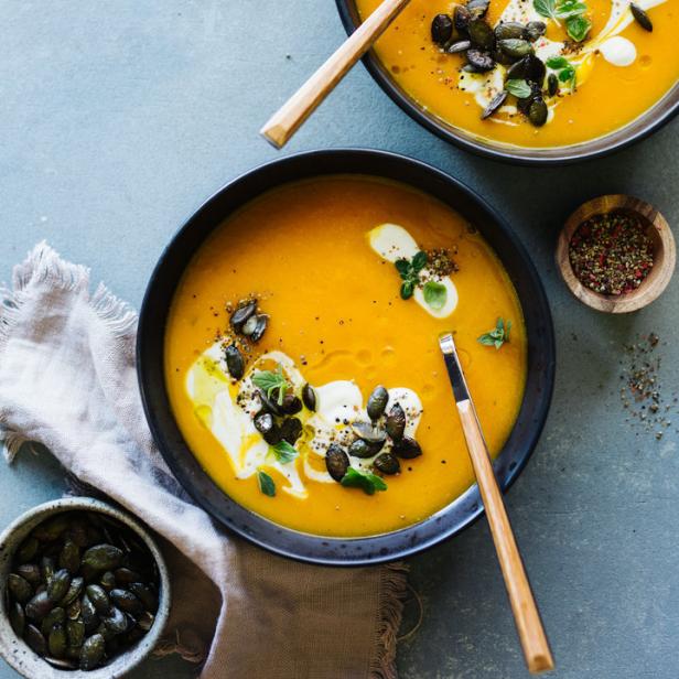 Zwei Schalen Kürbissuppe, garniert mit Kürbiskernen, Creme fraîche und Kräutern, stehen auf einem Tisch.