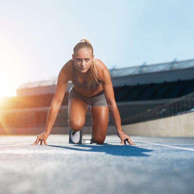 Eine blonde Frau in Sportkleidung kniet auf einer blauen Laufbahn in Startposition.